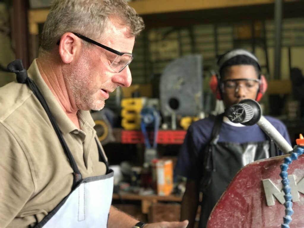 Artisan portant des lunettes de protection qui montre un travail à un jeune apprenti équipé d'un casque antibruit dans un atelier