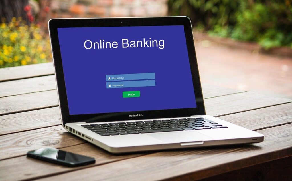 Ordinateur portable affichant une page de connexion de banque en ligne, smartphone posé à côté sur une table en bois