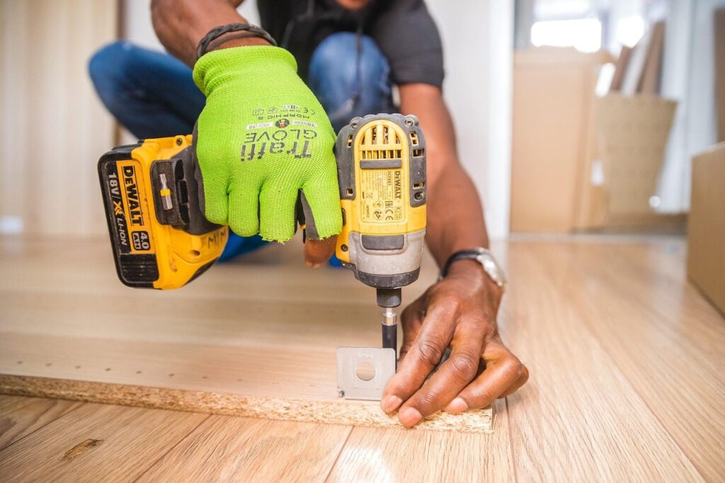 Artisan ganté assemblant un panneau de meuble avec une visseuse sans fil sur un parquet