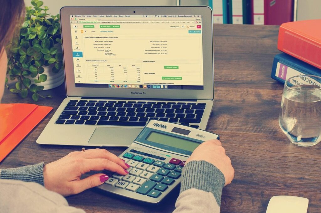 Personne utilisant une calculatrice devant un ordinateur portable affichant un logiciel de facturation et de comptabilité