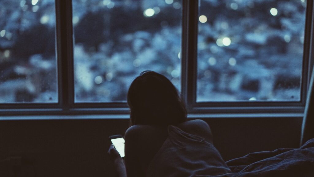 Personne allongée sur un lit, de dos, consultant son smartphone la nuit devant une fenêtre donnant sur les lumières de la ville